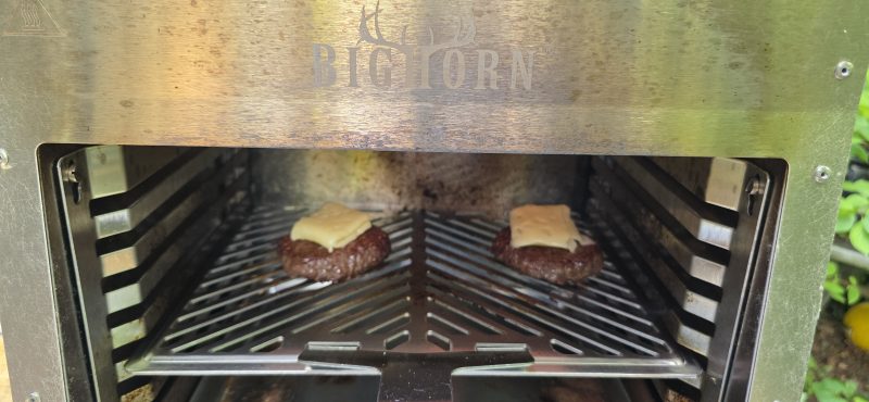 Two hamburger patties on the stainless rack of a Big Horn infrared broiler, each topped with a quartered piece of cheese starting to melt, with green foliage outside