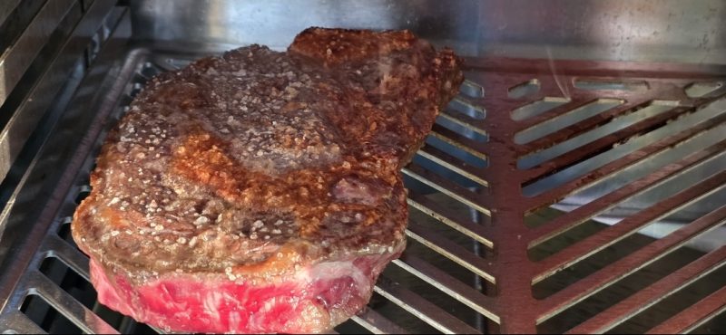 A ribeye steak searing on the stainless rack of a Big Horn infrared broiler, with a raw pink edge still showing