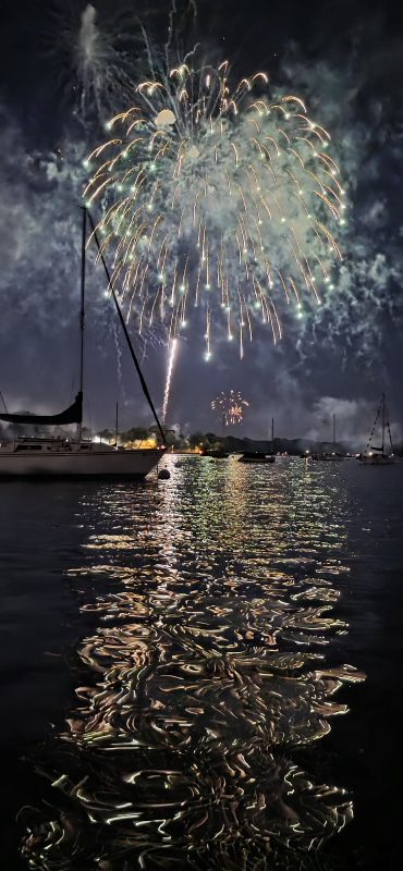 Fireworks reflecting on the calm harbor water at Neenah with sailboat silhouettes along the shore, Fourth of July night