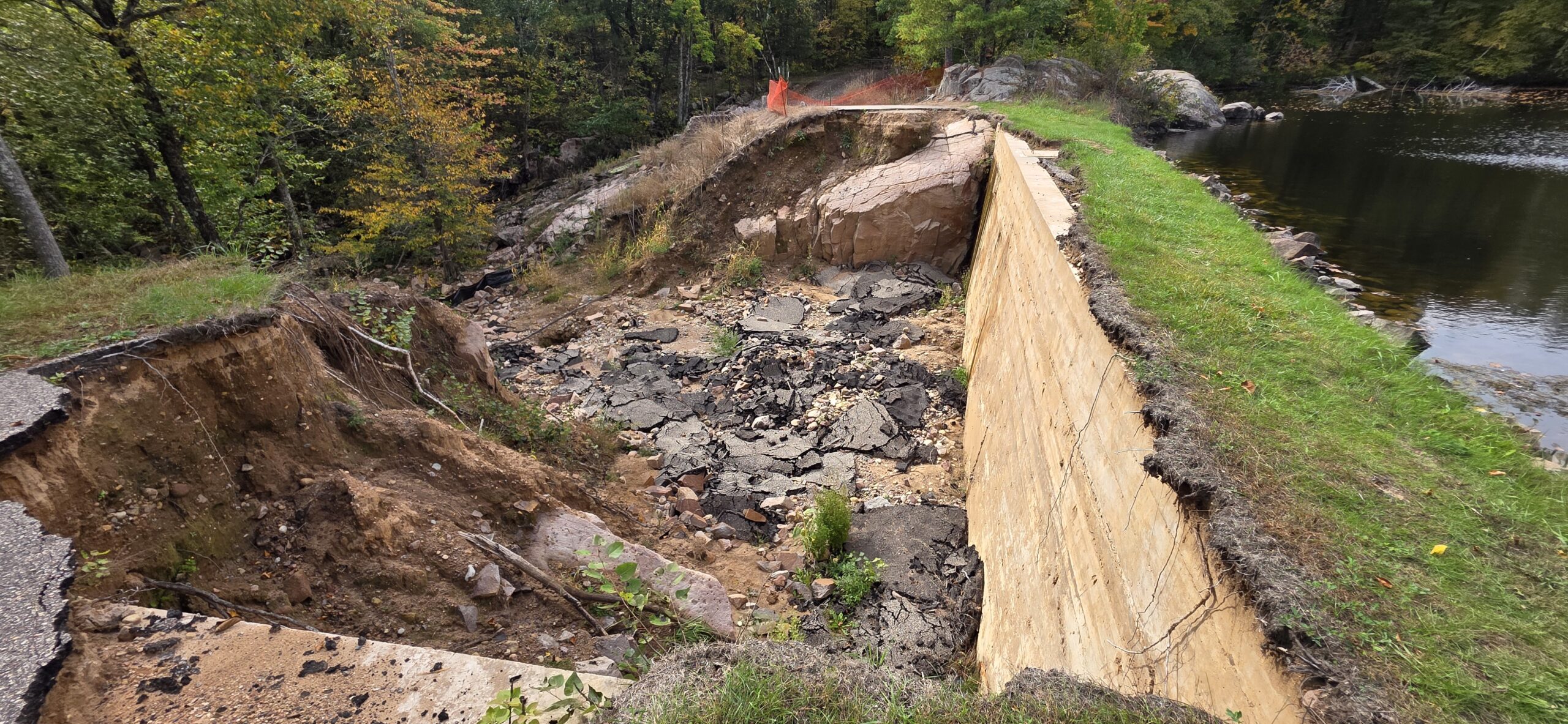 Severe washout damage showing a collapsed roadway and eroded earth next to a concrete retaining wall near a body of water.