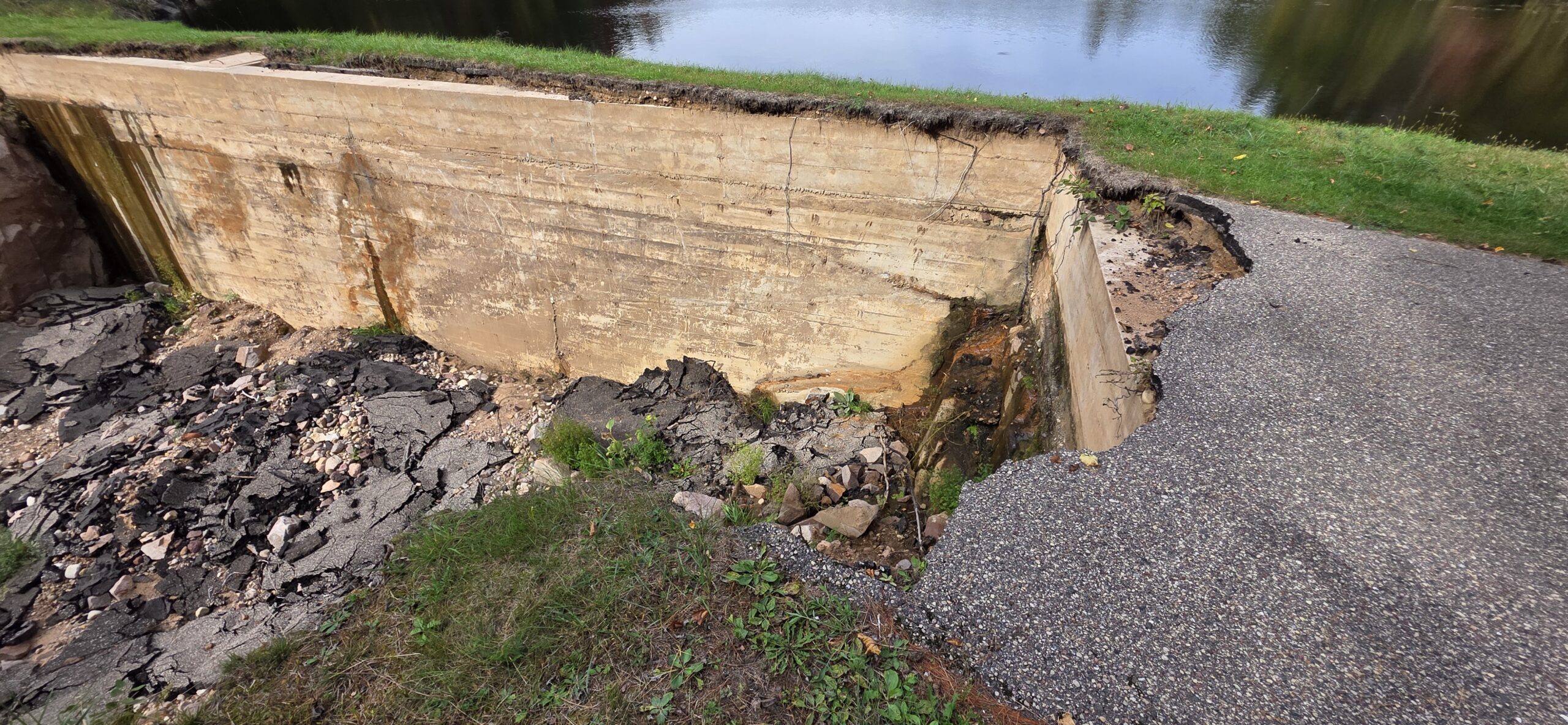 High-angle view of a damaged concrete dam structure with collapsed asphalt and eroded ground next to a body of water.