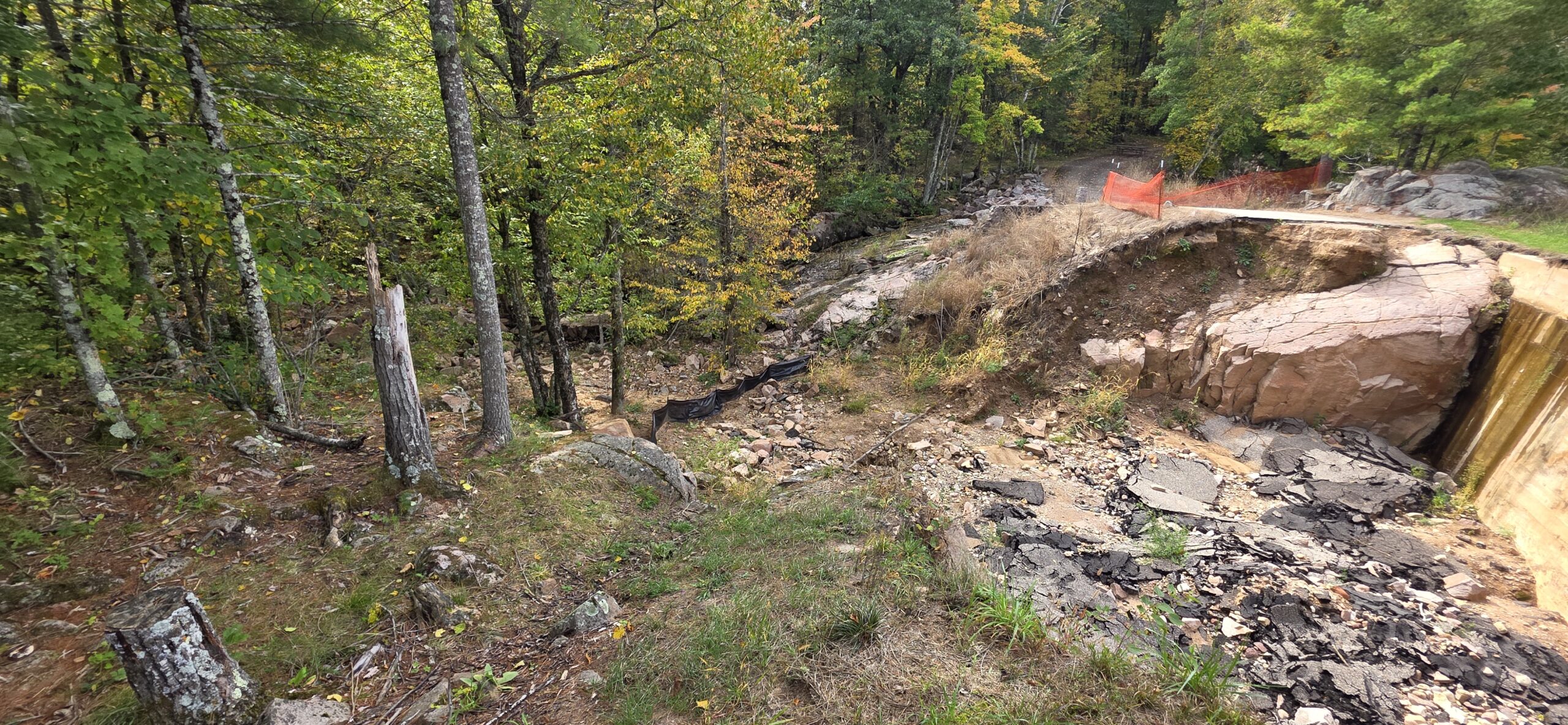 High angle view of a washed-out roadway next to a concrete retaining wall with broken asphalt and eroded soil.