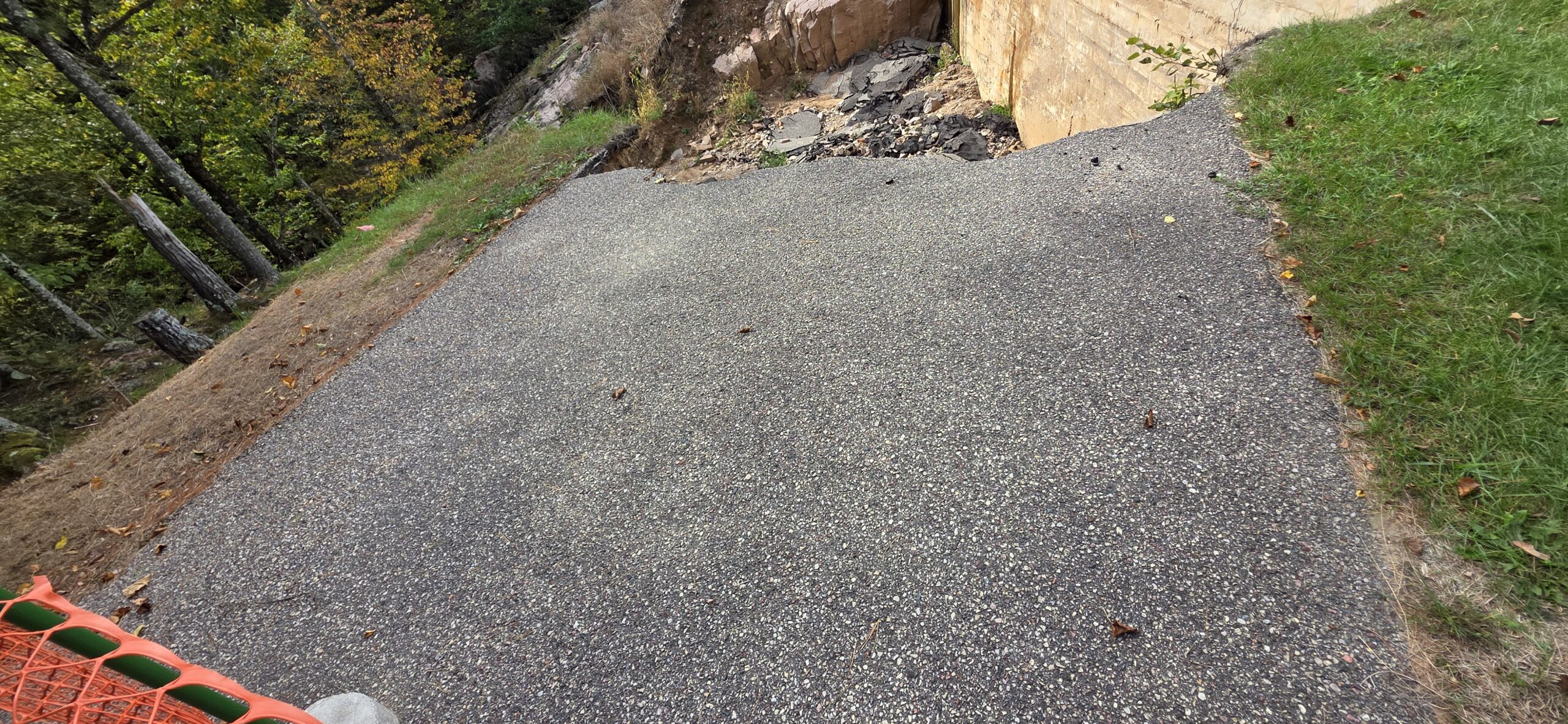 An elevated view showing a section of asphalt pavement ending abruptly at a washout near a concrete structure and rocky hillside.